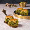 Three slices of baked baklava with a green pistachio-like filling on a round white plate, set on a dark marble surface with decorative objects in the background.