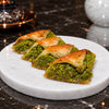 Three slices of baked baklava with a green pistachio-like filling on a round white plate, set on a dark marble surface with decorative objects in the background.
