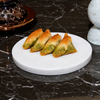 Four triangular pieces of golden-brown pastry with green filling arranged on a round white serving platter, set on a dark marble surface with decorative objects in the background.